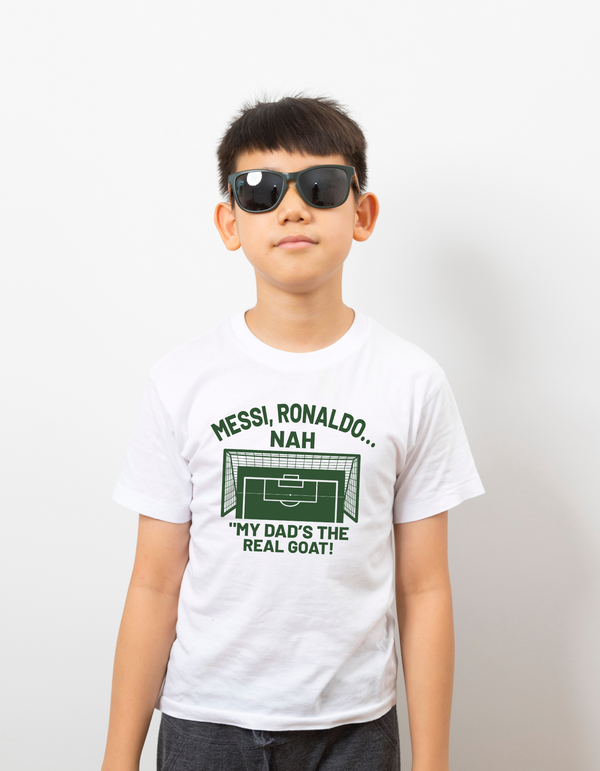 A little boy wearing a white t-shirt with the funny football and dad loving slogan on the front which reads 'Messi, Ronaldo…Nah ’My Dads the Real Goat!'