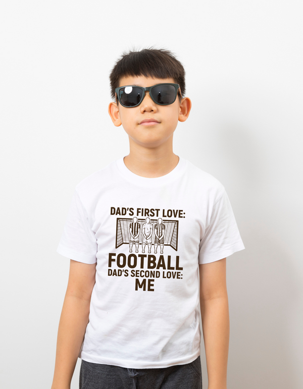 A boy wearing a white t-shirt with the funny football slogan on the front saying 'Dad’s First Love: Football. Dad’s Second Love: Me'