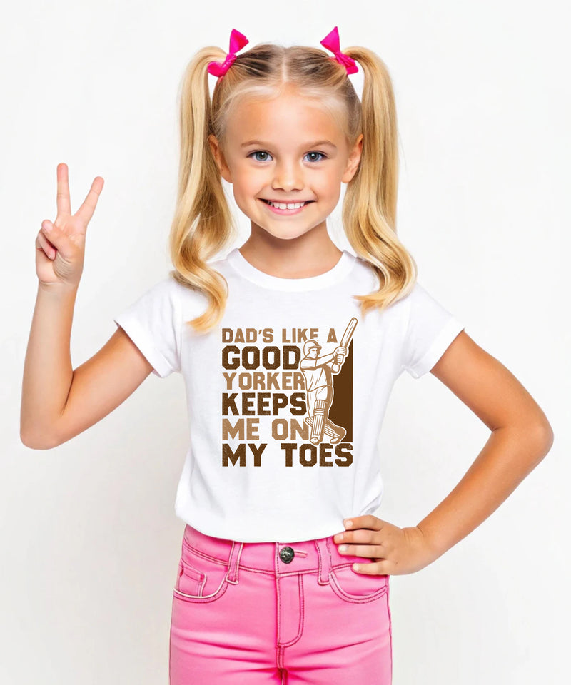 A little girl wearing a white t-shirt with Dads Like A Good Yorker, Keeps Me On My Toes on the front. A Funny Cricket T-Shirt, Premium Fabric, Multiple Junior Sizes, Perfect for Boys and Girls