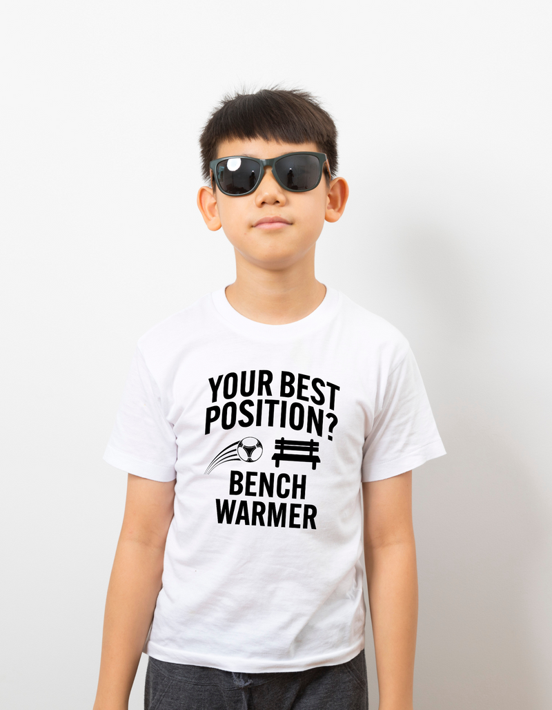 A little boy wearing a white t-shirt that has the funny football slogan on the front that reads 'Your Best Position? Bench Warmer'