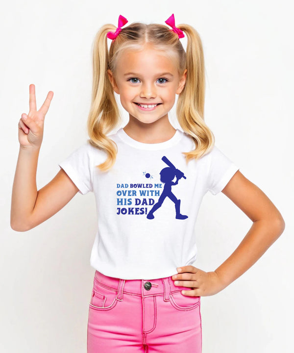 A little girl wearing a white t-shirt with Dad Bowled Me Over With His Dad Jokes on the front. A Funny Cricket T-shirt For Kids, Premium Fabric, Multiple Junior Sizes, Perfect For Boys And Girls