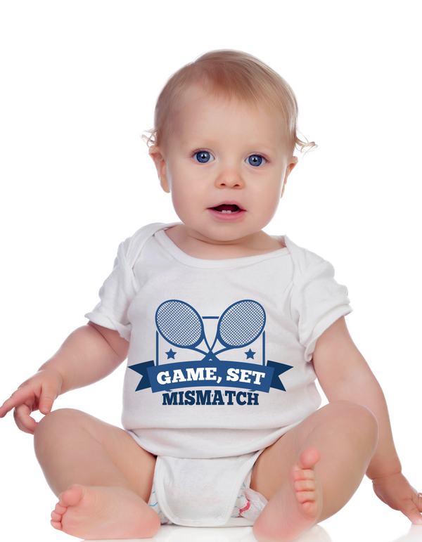A little baby wearing a white baby grow with the funny tennis slogan on the front that reads 'Game, Set, Mismatch'