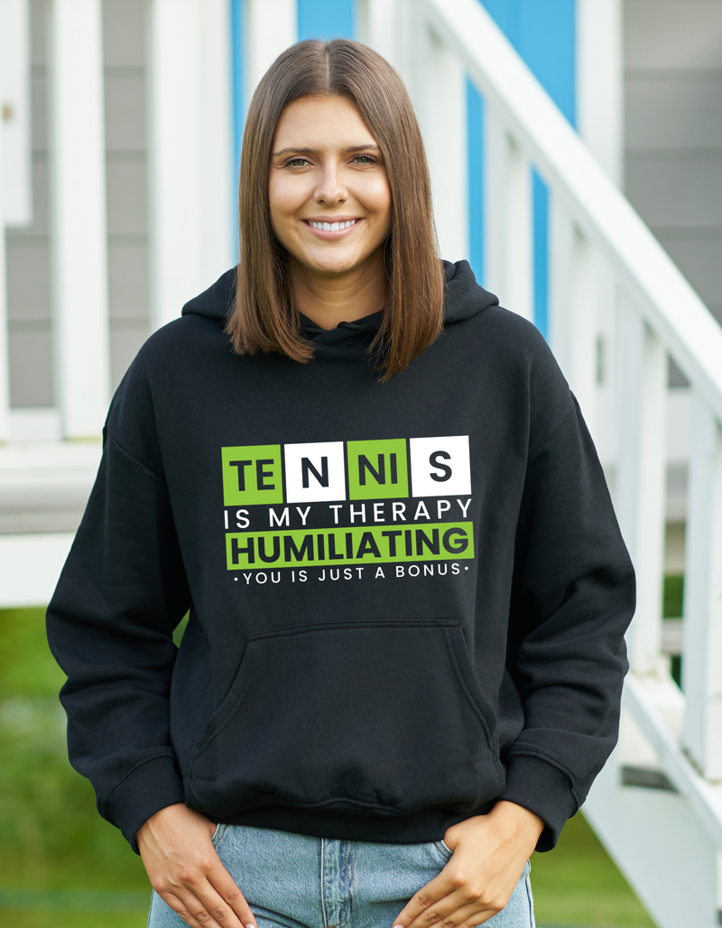 A lady wearing a black hoodie with the funny tennis slogan on the front that reads 'Tennis Is My Therapy, Humiliating You Is Just A Bonus'