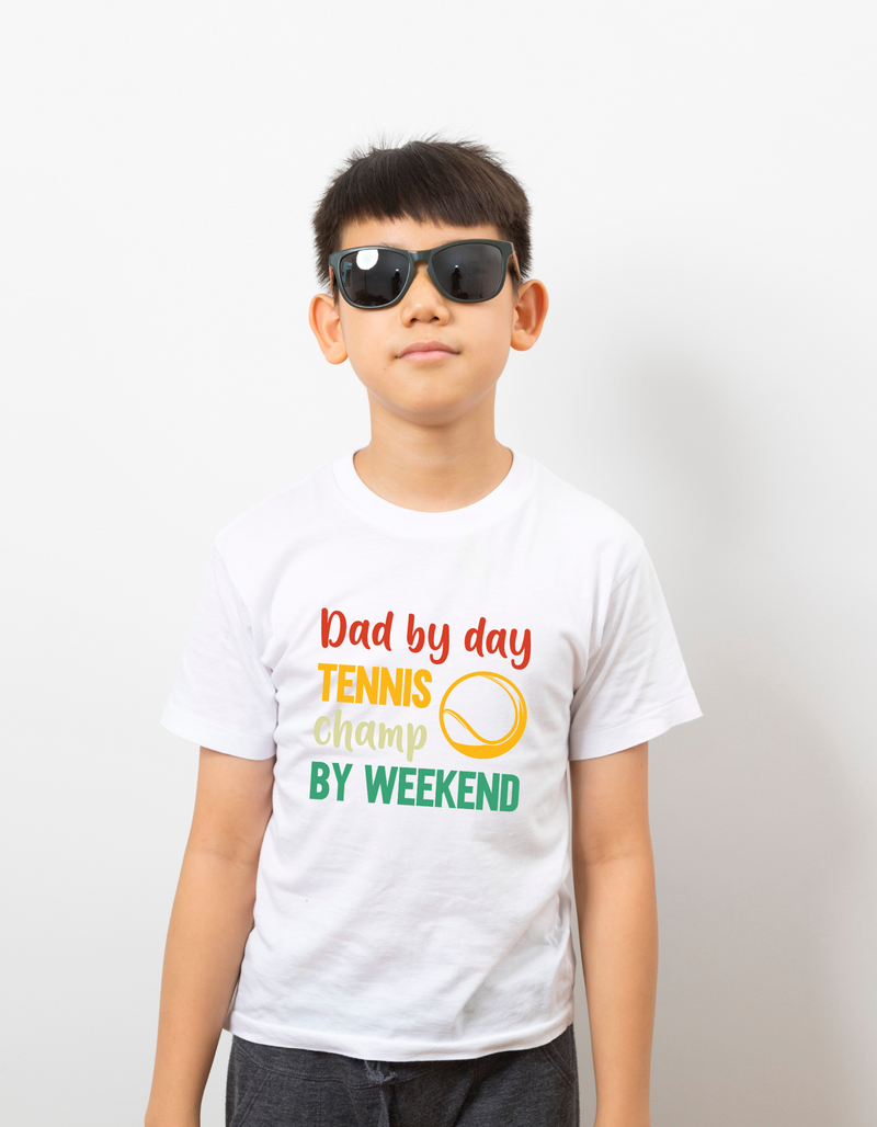 A boy wearing a white t-shirt with a funny tennis slogan on the front that reads 'Dad By Day, Tennis Champ By Weekend'