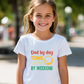 A girl wearing a white t-shirt with a funny tennis slogan on the front that reads 'Dad By Day, Tennis Champ By Weekend'