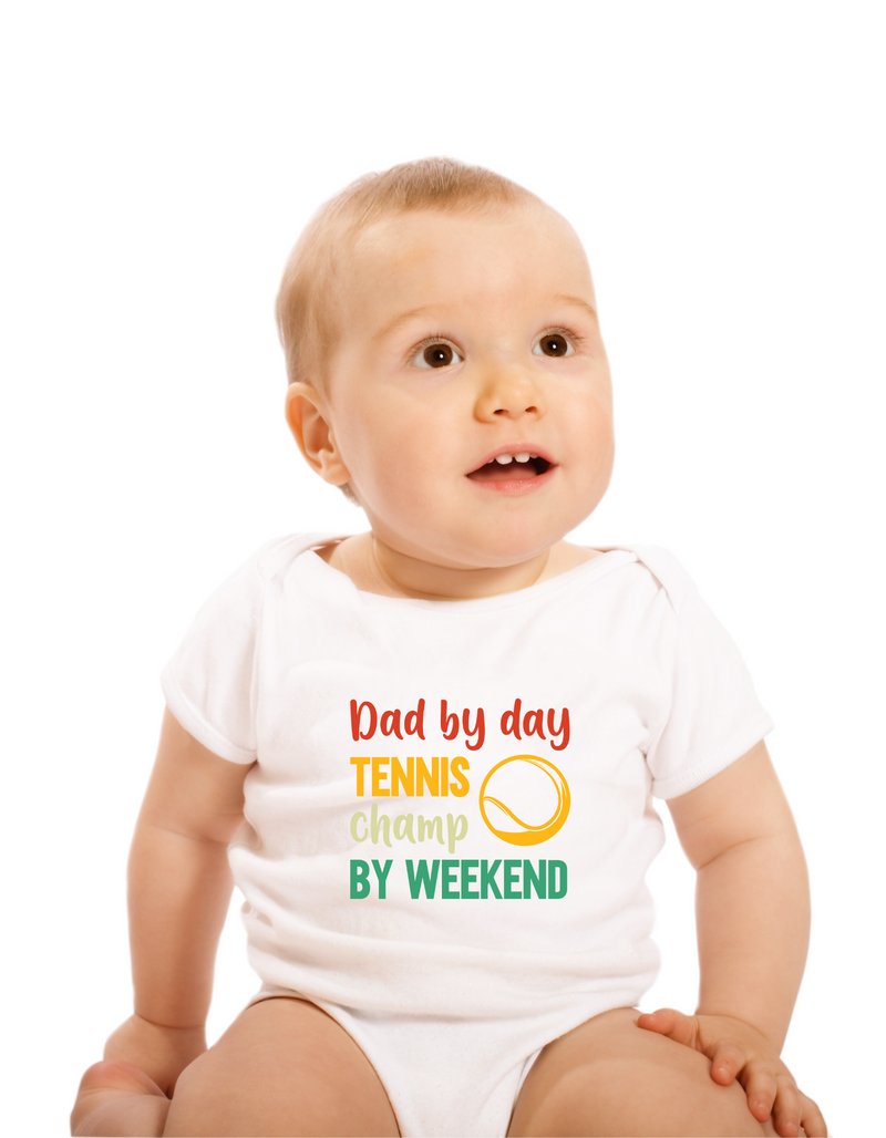 A little baby wearing a white baby grow with a funny tennis slogan on the front that reads 'Dad By Day, Tennis Champ By Weekend'
