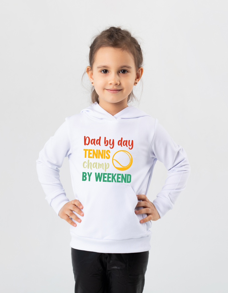 A little girl wearing a white hoodie with the funny tennis slogan on the front that reads 'Dad By Day, Tennis Champ By Weekend'