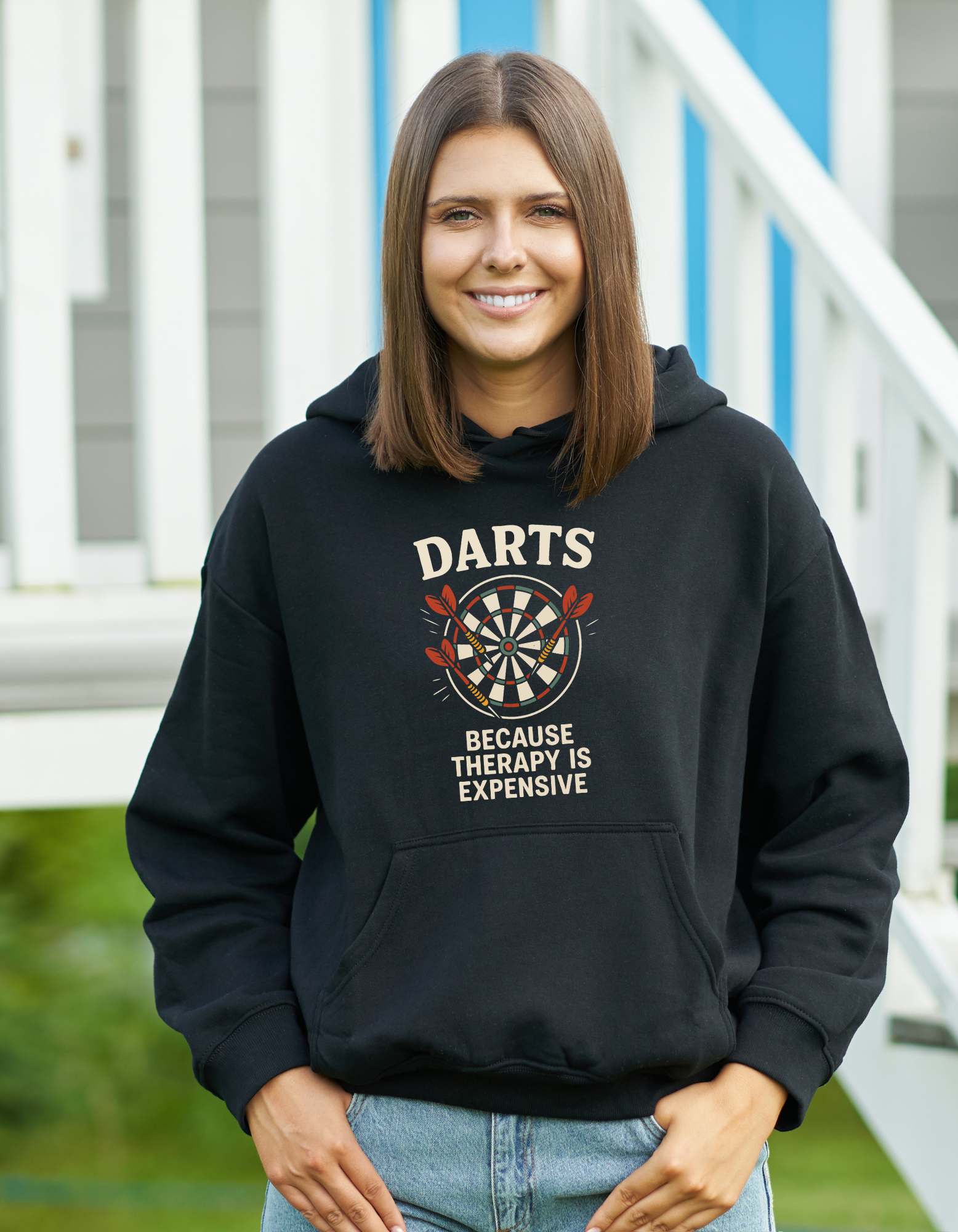 Woman wearing a black hoodie with 'Darts Because Therapy is Expensive' text and dartboard graphic with a Cows Corner slogan saying Darts because therapy is expensive.