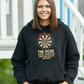 Woman wearing a black hoodie with a dartboard design and text, standing outdoors with a Cows Corner slogan The oche where legends are made and excuses are born.