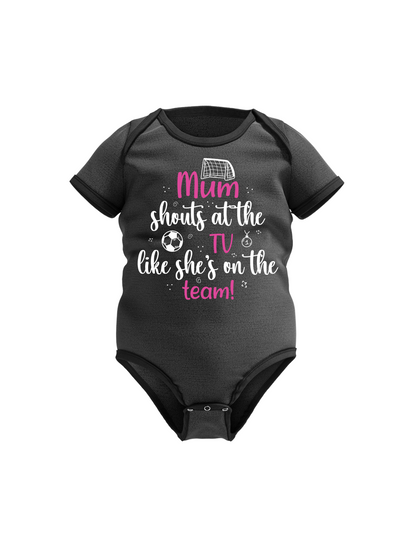 A black baby bodysuit with the text 'Mum Shouts at the TV Like she's on the Team!' along with graphic elements like a soccer ball a cows corner baby grow