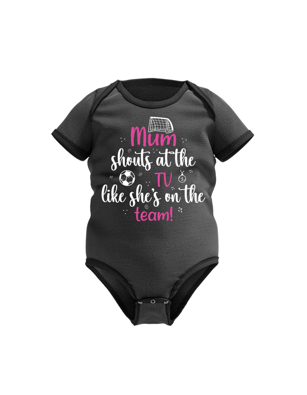 A black baby bodysuit with the text 'Mum Shouts at the TV Like she's on the Team!' along with graphic elements like a soccer ball a cows corner baby grow