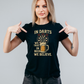 Woman wearing a black t-shirt with a dart and beer graphic, pointing at the text on a gray background with a Cows Corner slogan In darts we trust, in beer we believe.