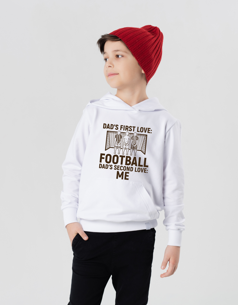 A young boy wearing a white hoodie with the funny football slogan on the front saying 'Dad’s First Love: Football. Dad’s Second Love: Me'