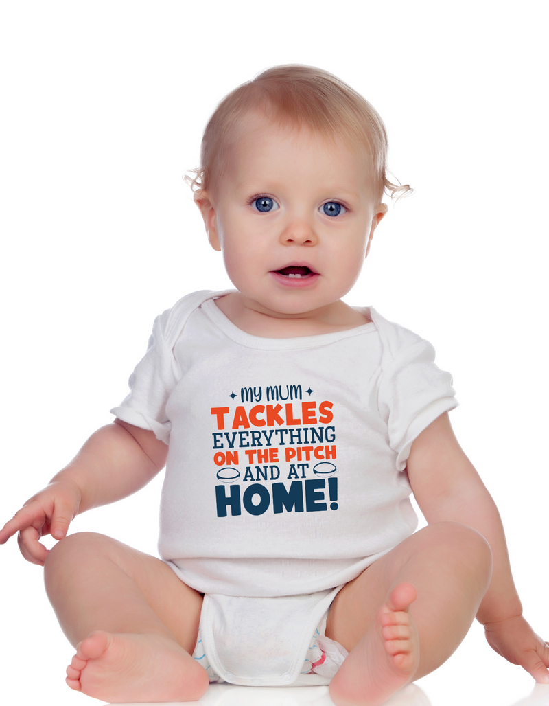 Baby wearing a white onesie with colorful text on a white background, with a slogan on the front saying 'My Mum Tackles Everything on the Pitch and at Home!'