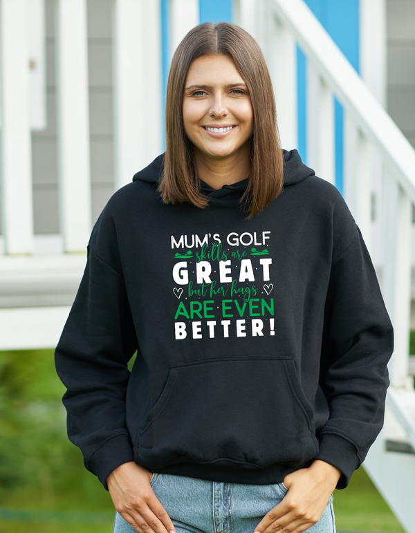 Woman wearing a black hoodie with humorous text, standing outdoors with a Cows Corner slogan saying Mum's golf skills are great, but her hugs are even better.
