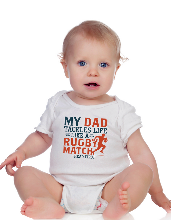 Baby wearing a white onesie with a colourful text design on a white background with the Cows Corner slogan saying My dad tackles life like a rugby match - head first. 