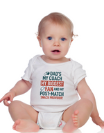 Baby wearing a white onesie with text on a white background, with a slogan on the front saying 'Dad’s My Coach My Biggest Fan and My Post-Match Snack Provider!