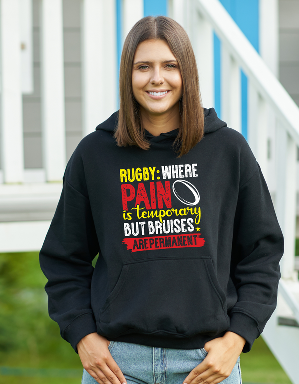 Woman wearing a black hoodie with a colourful text design outdoors with a Cows Corner slogan saying Rugby where pain is temporay but bruises are permanent