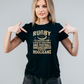 Woman wearing a black t-shirt with rugby-themed text, pointing at the shirt, with slogan on the front saying 'Rugby Is a Hooligan’s Game Played by Gentleman, and Football Is a Gentleman’s Game Played by Hooligan
