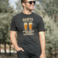 Man wearing a black t-shirt with a humorous dart and beer graphic, standing outdoors with a Cows Corner slogan saying Darts the only sport where beeer improves performance.