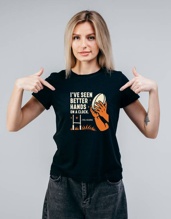 Woman wearing a black t-shirt with a graphic and text, pointing at the shirt, slogan saying 'I’ve Seen Better Hands on a Clock – Phil Kearns'