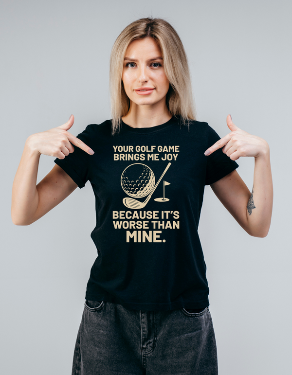 Woman wearing a black t-shirt with a humorous golf-themed design, pointing at the text with Cows Corner slogan saying, Your golf game brings me joy, because it's worse than mine
