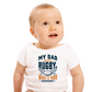 Baby wearing a white onesie with text about rugby, sitting on a white background, with a slogan on the front saying 'My Dad Plays Rugby. What’s Your Superpower?