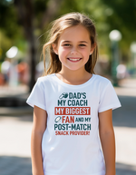 Young girl wearing a white t-shirt with a humorous message, standing outdoors, with a slogan on the front saying 'Dad’s My Coach My Biggest Fan and My Post-Match Snack Provider!