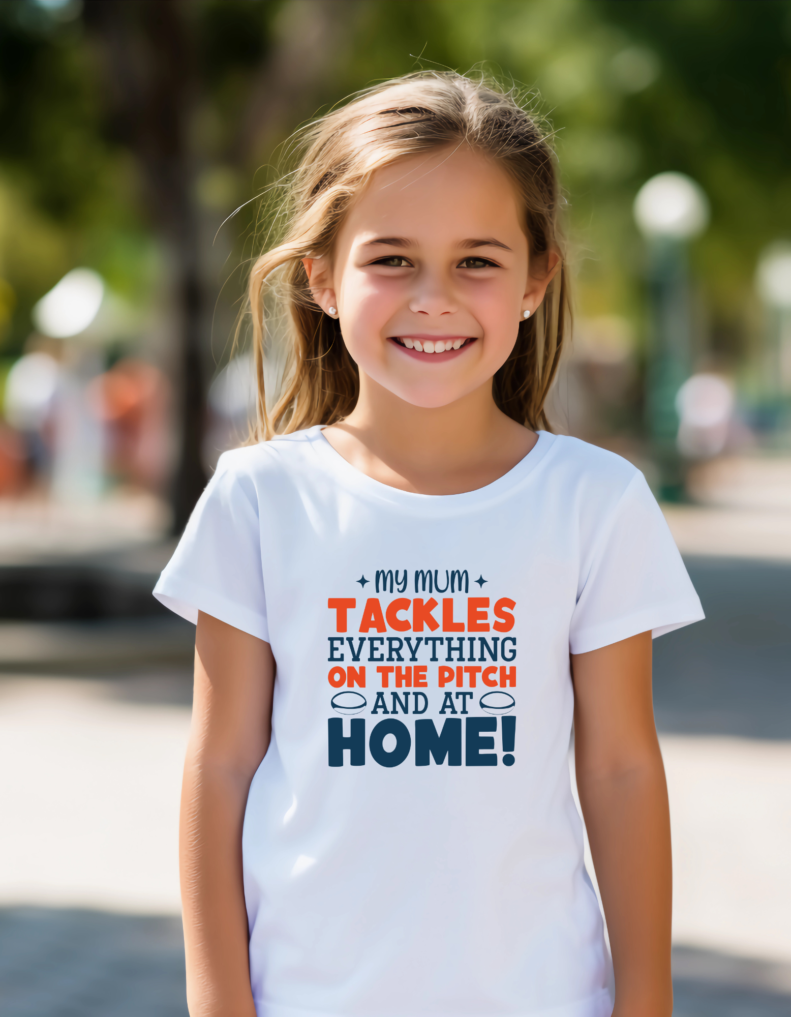 Young girl wearing a white t-shirt with colorful text in an outdoor setting, with a slogan on the front saying 'My Mum Tackles Everything on the Pitch and at Home!'