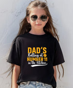 A little girl wearing a black t-shirt with Dads Batting At Number 11 In The Kitchen on the front of the T-Shirt. Funny Cricket T-shirt, Premium Fabric, Multiple Junior Sizes, Perfect for Boys and Girls