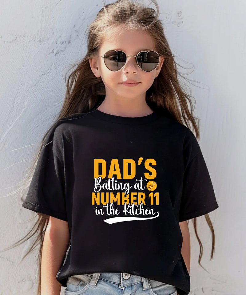 A little girl wearing a black t-shirt with Dads Batting At Number 11 In The Kitchen on the front of the T-Shirt. Funny Cricket T-shirt, Premium Fabric, Multiple Junior Sizes, Perfect for Boys and Girls