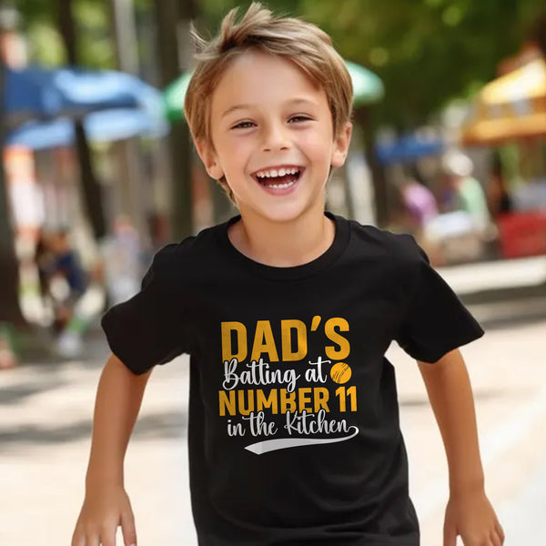 A little boy wearing a black t-shirt with Dads Batting At Number 11 In The Kitchen on the front of the T-Shirt. Funny Cricket T-shirt, Premium Fabric, Multiple Junior Sizes, Perfect for Boys and Girls