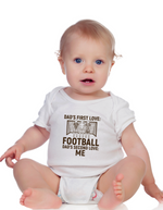 A baby wearing a white baby grow with the funny football slogan on the front saying 'Dad’s First Love: Football. Dad’s Second Love: Me'