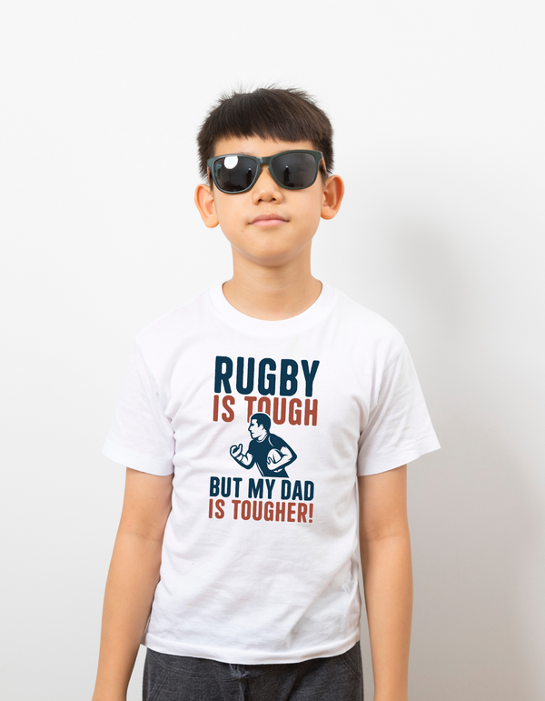 Child wearing a white t-shirt with rugby-themed text and graphics on a plain background with a Cows Corner slogan saying Rugby is tough, but my dad is tougher 