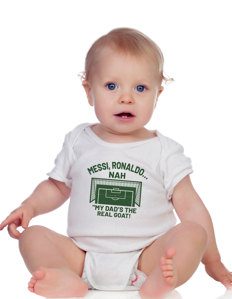 A little baby wearing a white baby grow with the funny football and dad loving slogan on the front which reads 'Messi, Ronaldo…Nah ’My Dads the Real Goat!'