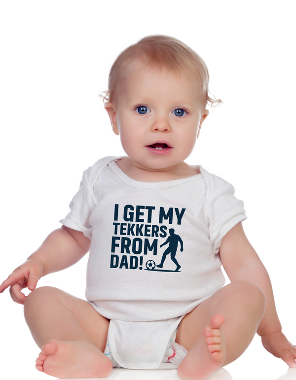 A little baby wearing a baby grow with the funny football slogan on the front saying 'I Get My Tekkers From Dad'