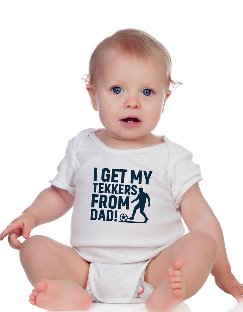 A little baby wearing a baby grow with the funny football slogan on the front saying 'I Get My Tekkers From Dad'