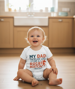 Baby sitting on a wooden floor wearing a onesie with a humorous message.  A Cows corner baby grow with slogan 'My Dad Tackles Life Like a Rugby Match- Head First'. Cows Corner gifts are perfect for sport-mad fans, these gifts work brilliantly for birthdays, new baby celebrations, Father’s Day, Mother’s Day, Christmas, anniversaries, thank you gifts, end-of-season team awards, graduations, retirements, and just-because moments when you want to raise a smile. 
