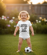 Child wearing a onesie with a humorous message, standing on grass with a soccer ball. The baby is wearing a Cows Corner baby grow with the funny football slogan 'Your Best Position? Bench Warmer'.  Cows Corner gifts are perfect for sport-mad fans, these gifts work brilliantly for birthdays, new baby celebrations, Father’s Day, Mother’s Day, Christmas, anniversaries, thank you gifts, end-of-season team awards, graduations, retirements, and just-because moments when you want to raise a smile. 
