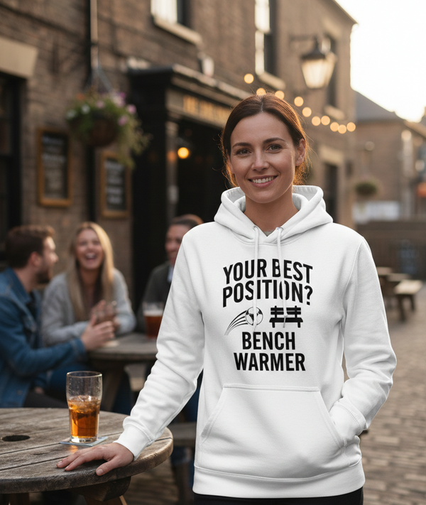 Woman wearing a white hoodie with text, standing outdoors in a pub setting. The lady is wearing a Cows Corner hoodie with a funny football slogan 'Your Best Position? Bench Warmer'. Cows Corner gifts are perfect for sport-mad fans, these gifts work brilliantly for birthdays, new baby celebrations, Father’s Day, Mother’s Day, Christmas, anniversaries, thank you gifts, end-of-season team awards, graduations, retirements, and just-because moments when you want to raise a smile. 

