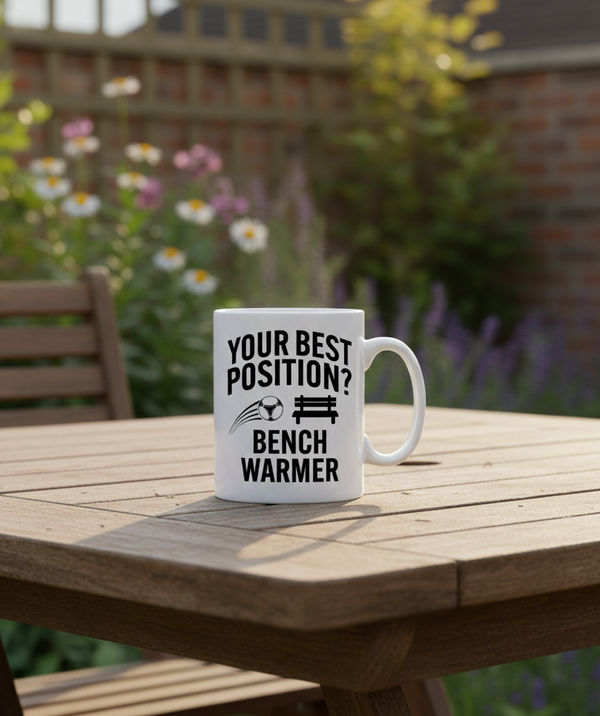White mug with humorous text on a wooden table outdoors. This is a Cows Corner mug with a funny football slogan 'Your Best Position? Bench Warmer'. Cows Corner gifts are perfect for sport-mad fans, these gifts work brilliantly for birthdays, new baby celebrations, Father’s Day, Mother’s Day, Christmas, anniversaries, thank you gifts, end-of-season team awards, graduations, retirements, and just-because moments when you want to raise a smile. 
