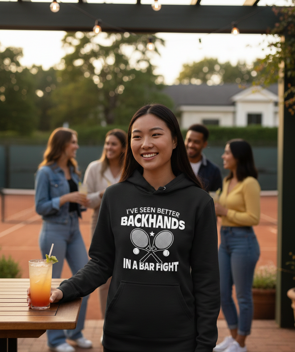 Woman wearing a black hoodie with a humorous tennis graphic and text, standing outdoors with friends in the background.  The lady is wearing a Cows Corner hoodie with a funny tennis slogan 'I've Seen Better Backhands In A Bar Fight'. Cows Corner gifts are perfect for sport-mad fans, these gifts work brilliantly for birthdays, new baby celebrations, Father’s Day, Mother’s Day, Christmas, anniversaries, thank you gifts, end-of-season team awards, graduations, retirements, and just-because moments when you wan