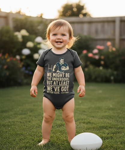 Child wearing a onesie with text and a graphic, standing outdoors on grass. The baby is wearing a Cows Corner baby grow with a funny rugby slogan 'We Might Be the Underdogs, but at Least We’ve Got Better Teeth – Scott Gibbs'. Cows Corner gifts are perfect for sport-mad fans, these gifts work brilliantly for birthdays, new baby celebrations, Father’s Day, Mother’s Day, Christmas, anniversaries, thank you gifts, end-of-season team awards, graduations, retirements, and just-because moments 