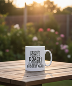 White mug with text and graphic on a wooden table outdoors. This is a Cows Corner mug with a funny football slogan 'Dad’s My Coach My Hero and My Biggest Fan!'. Cows Corner gifts are perfect for sport-mad fans, these gifts work brilliantly for birthdays, new baby celebrations, Father’s Day, Mother’s Day, Christmas, anniversaries, thank you gifts, end-of-season team awards, graduations, retirements, and just-because moments when you want to raise a smile. 

