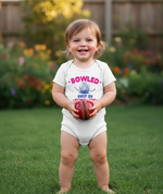 Child holding a red ball in a garden wearing a onesie with text. The baby is wearing a Cows Corner baby grow with a funny cricket slogan 'Bowled Over by My Mum’s Love & Cooking'. Cows Corner gifts are perfect for sport-mad fans, these gifts work brilliantly for birthdays, new baby celebrations, Father’s Day, Mother’s Day, Christmas, anniversaries, thank you gifts, end-of-season team awards, graduations, retirements, and just-because moments when you want to raise a smile. 
