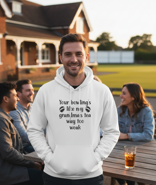 Man wearing a white hoodie with text, standing outdoors with friends at a table. The man is wearing a Cows Corner hoodie with a funny cricket slogan 'Your bowling is like my grandma's tea, way too weak!'.  Cows Corner gifts are perfect for sport-mad fans, these gifts work brilliantly for birthdays, new baby celebrations, Father’s Day, Mother’s Day, Christmas, anniversaries, thank you gifts, end-of-season team awards, graduations, retirements, and just-because moments when you want to raise a smile. 
