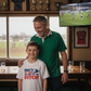 Man and young boy standing together in a pub with a TV showing a sports match. The boy is wearing a Cows Corner t-shirt with the funny rugby slogan on the front that reads 'Dad’s My Hero on and off the Pitch!'. Cows Corner gifts are perfect for sport-mad fans, these gifts work brilliantly for birthdays, new baby celebrations, Father’s Day, Mother’s Day, Christmas, anniversaries, thank you gifts, end-of-season team awards, graduations, retirements, and just-because moments when you want to raise a smile. 
