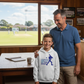 Man and child standing in a room with a view of a baseball field, the boy is wearing a Cows Corner hoodie with the funny cricket slogan 'Dad Bowled Me Over With His Dad Jokes'. Cows Corner gifts are perfect for sport-mad fans, these gifts work brilliantly for birthdays, new baby celebrations, Father’s Day, Mother’s Day, Christmas, anniversaries, thank you gifts, end-of-season team awards, graduations, retirements, and just-because moments when you want to raise a smile. 
