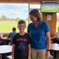 Woman and child standing together in a sports bar with a TV showing a cricket match.  The young boy is wearing a Cows Corner t-shirt that has a funny and sweet cricket slogan on the front that reads 'Bowled Over by My Mum’s Love & Cooking'