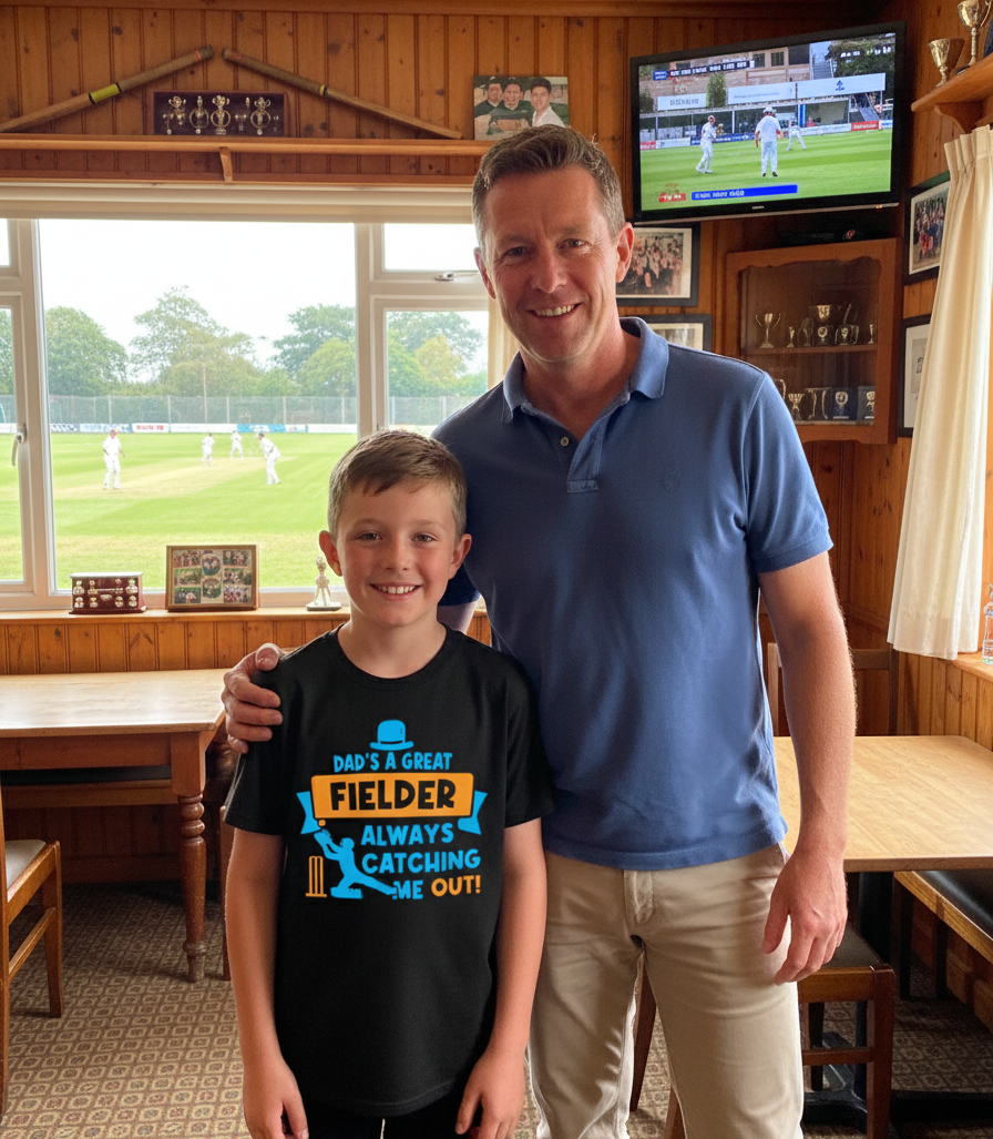 Man and young boy standing together in a room with a sports game on TV.  The young boy is wearing a Cows Corner t-shirt with the funny and sweet cricket slogan on the front that reads 'Dad is a Great Fielder, Always Catching Me Out'